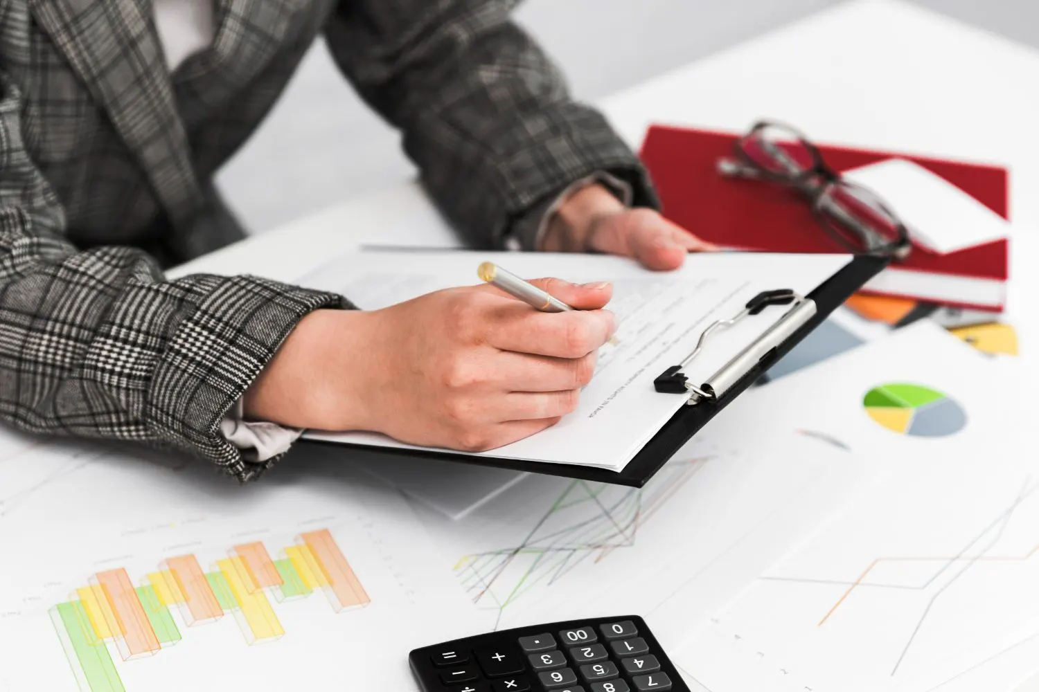 Mujer escribiendo en un portapapeles sobre un escritorio con gráficos y una calculadora