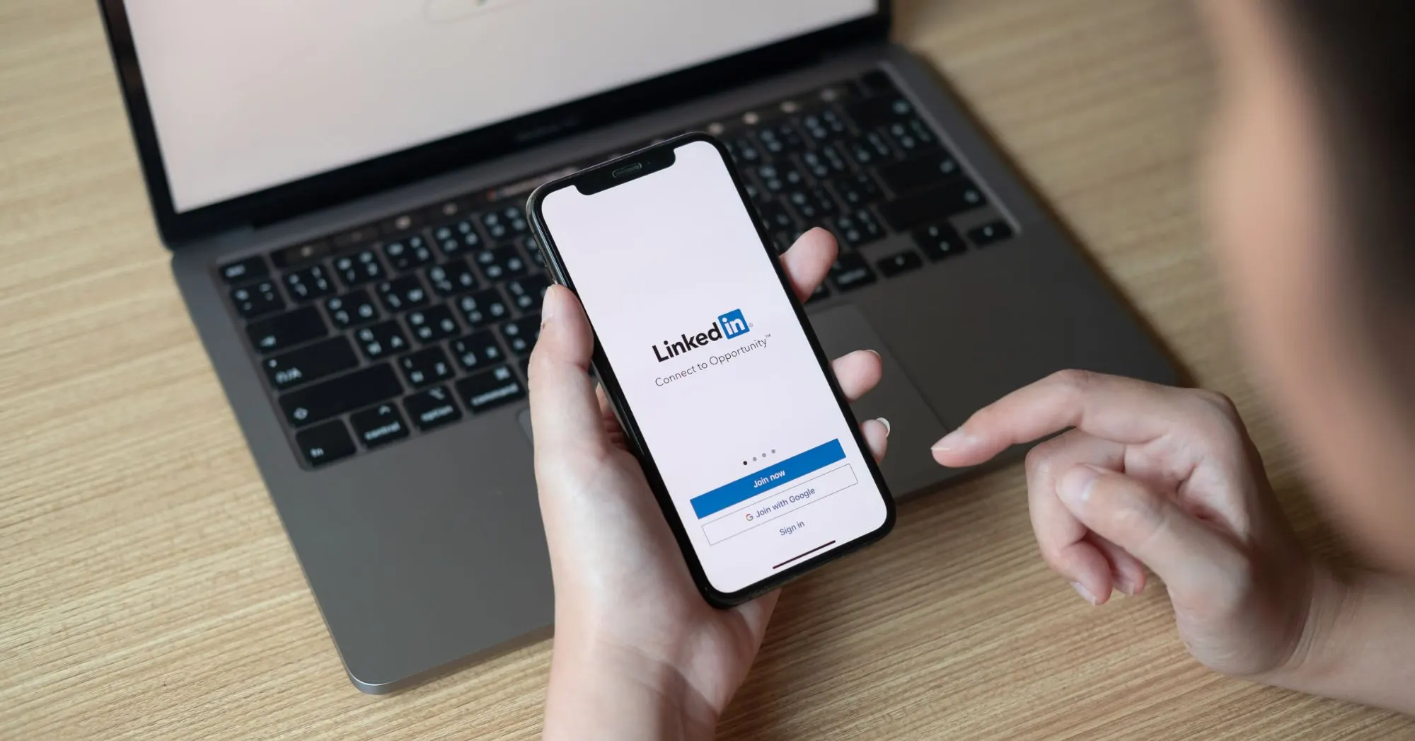 Manos con un móvil que muestra la pantalla de inicio de LinkedIn delante de un portátil