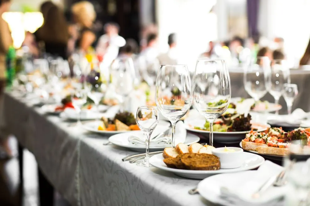Taula de banquet amb copes, pa i plats servits en un esdeveniment amb convidats al fons