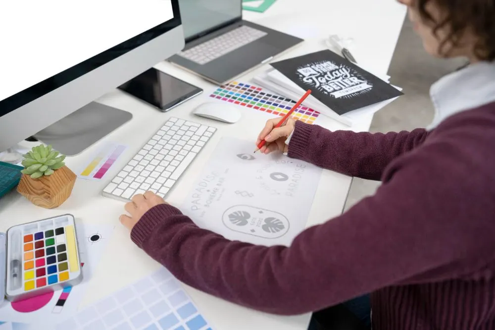 Diseñadora bocetando un logotipo a lápiz en un escritorio con ordenador, carta de colores y acuarelas
