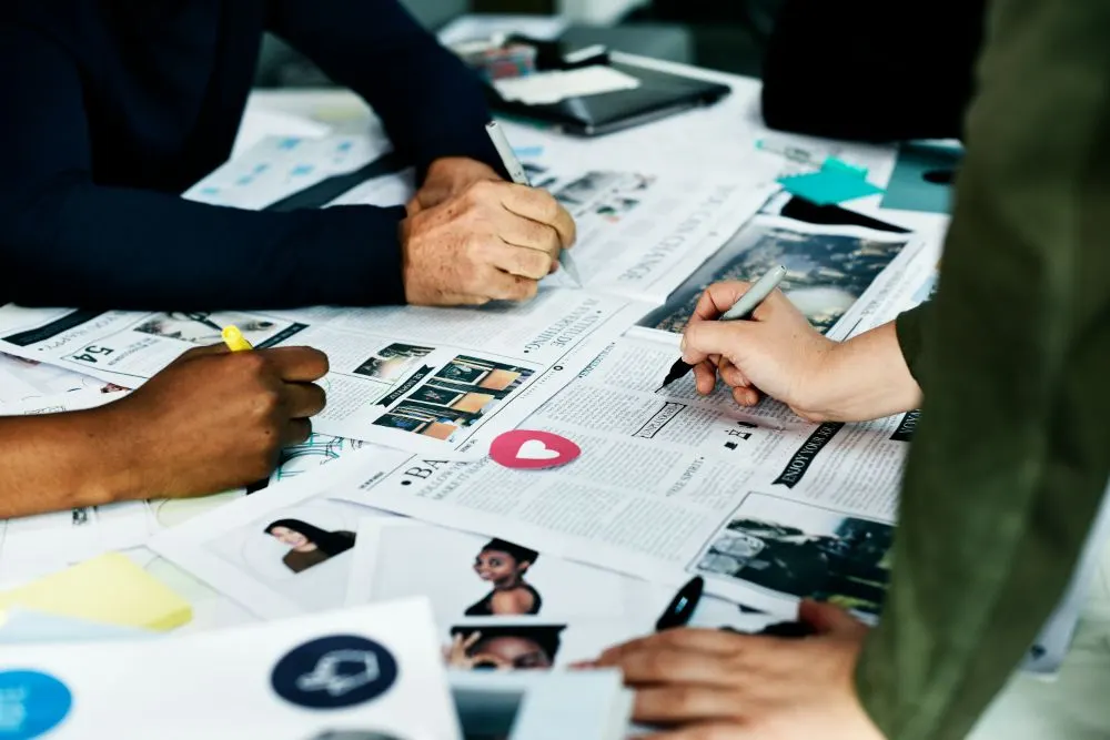 Manos de varias personas revisando y marcando revistas y recortes impresos sobre una mesa