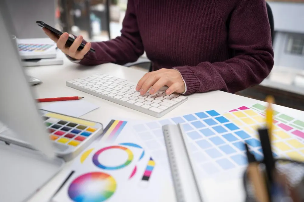 Diseñadora con móvil y teclado junto a paletas de colores, círculo cromático y una regla