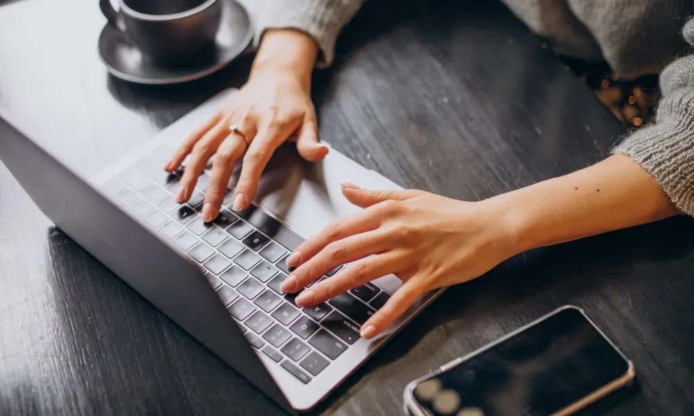 Manos escribiendo en un portátil sobre una mesa oscura junto a una taza de café y un móvil