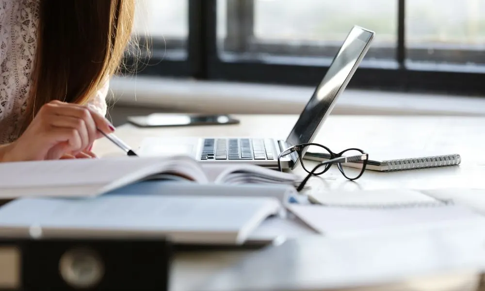 Mujer tomando notas junto a un portátil, libros abiertos y unas gafas sobre la mesa