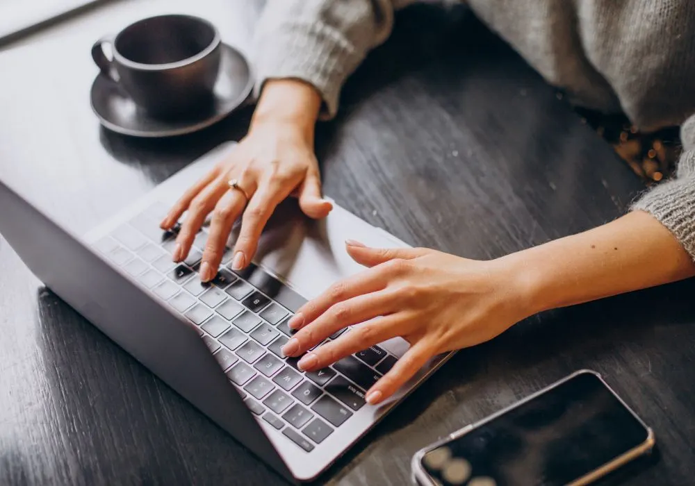 Manos escribiendo en un portátil junto a una taza de café y un móvil