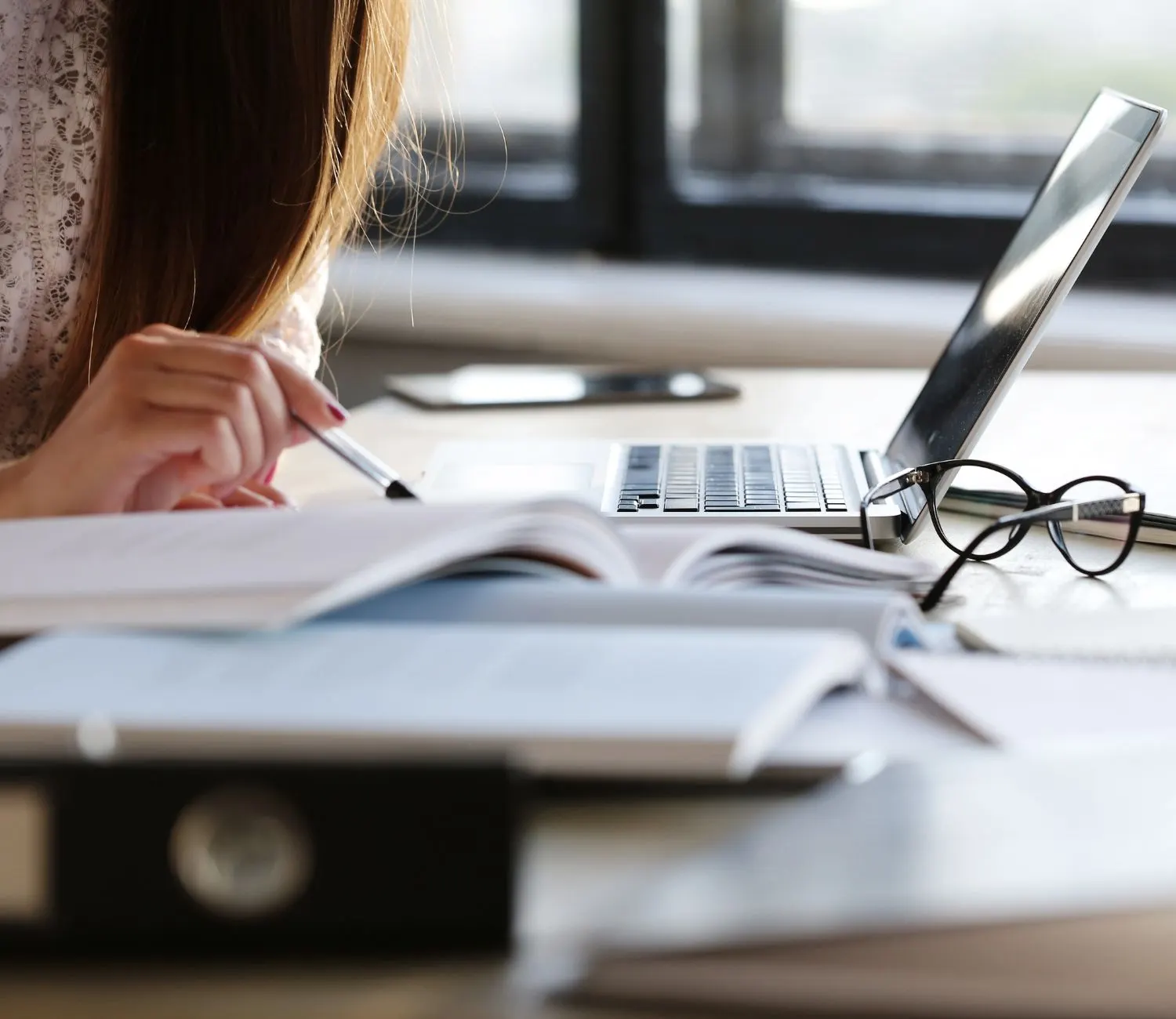 Mujer tomando notas junto a un portátil, libros abiertos y unas gafas sobre la mesa