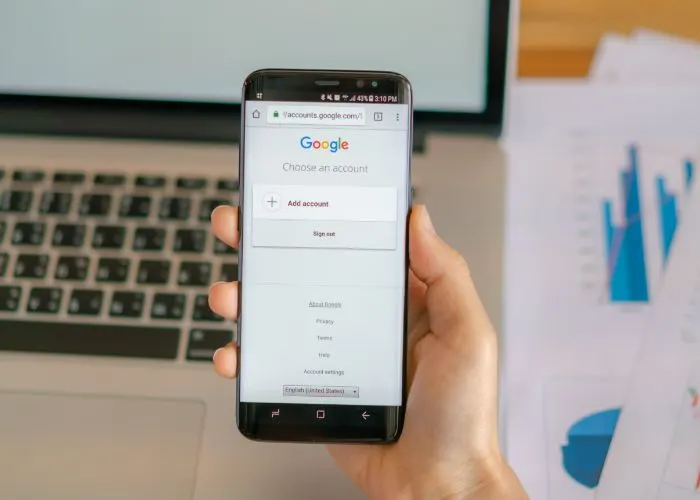 Mà sostenint un mòbil amb la pantalla d’inici de sessió de Google al costat d'un portàtil