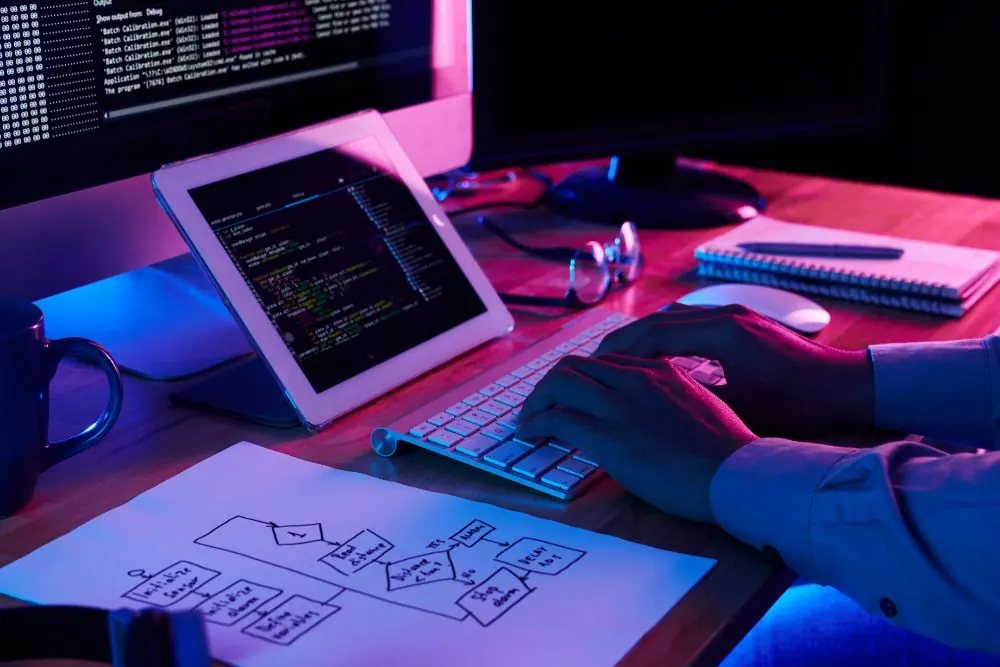 Manos tecleando junto a una tablet con código y un diagrama de flujo en papel, con luz de neón