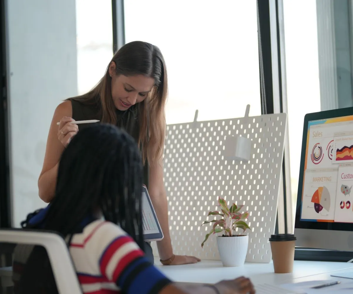 Dos mujeres trabajando en una oficina junto a un monitor con gráficos de ventas y marketing
