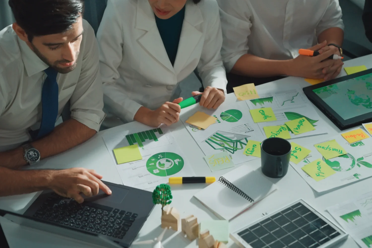 Equipo trabajando con gráficos de sostenibilidad, notas adhesivas, portátil y un panel solar en miniatura