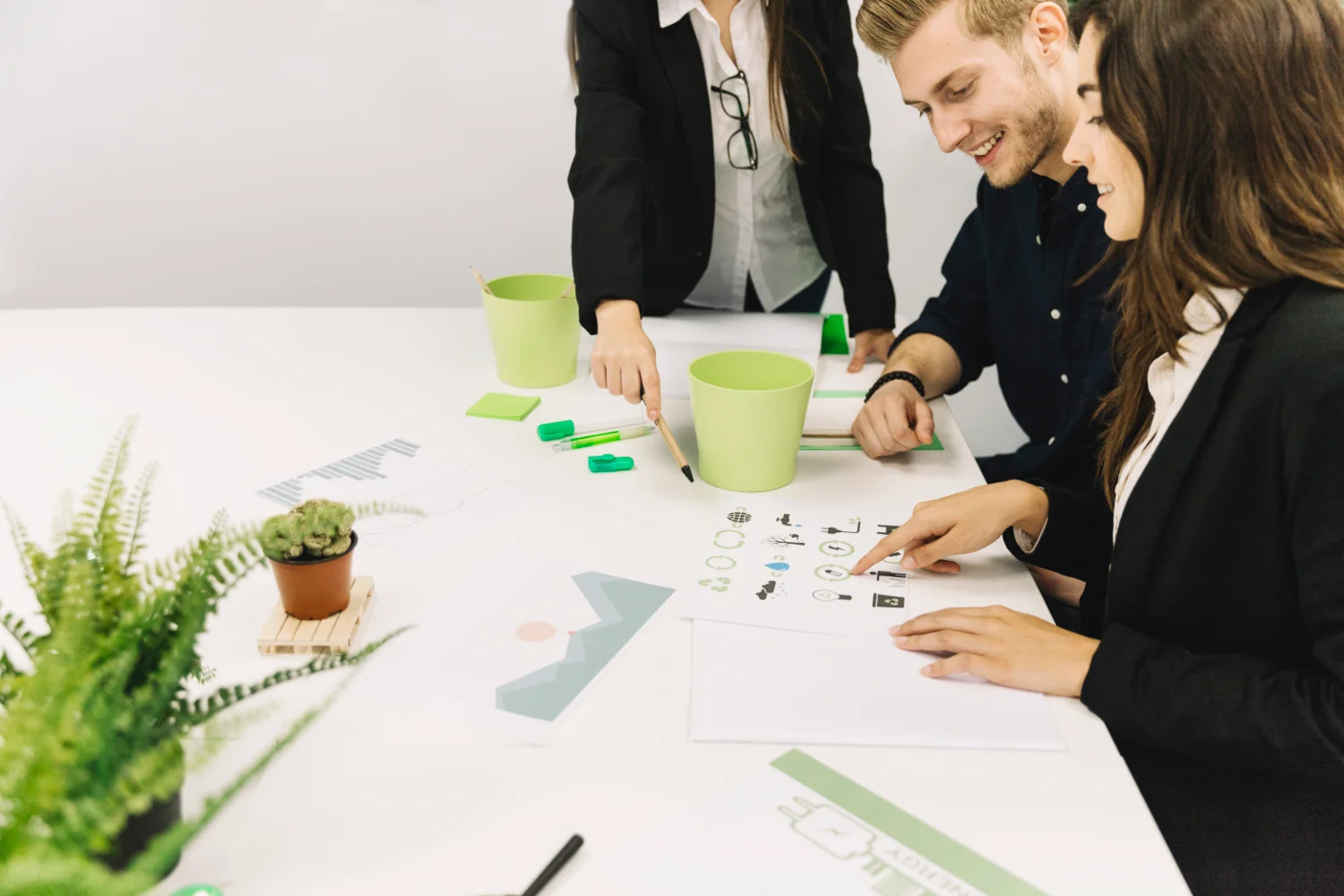 Tres compañeros sonrientes revisando iconos ecológicos impresos sobre una mesa con plantas