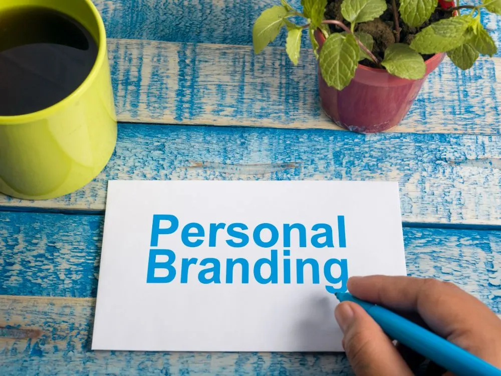 Mano escribiendo «Personal Branding» en una tarjeta sobre una mesa azul con taza y planta