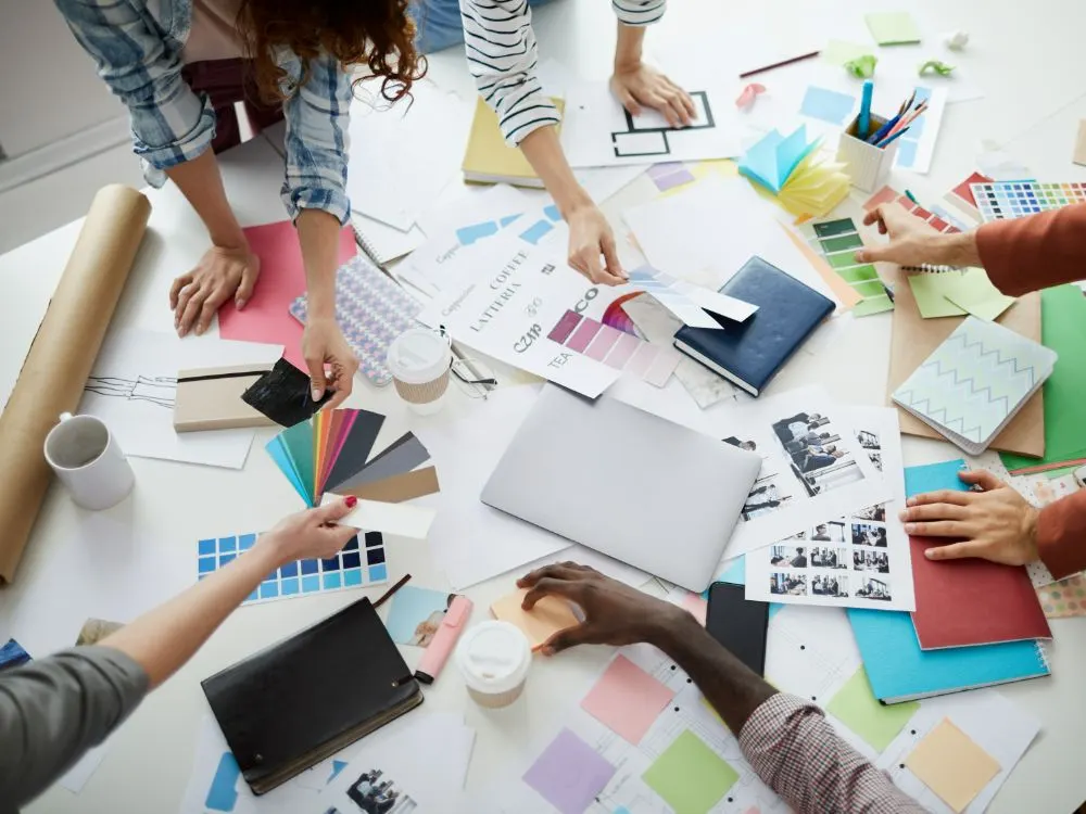 Vista zenital d’un equip creatiu amb mostres de color, esbossos i fotos sobre una taula