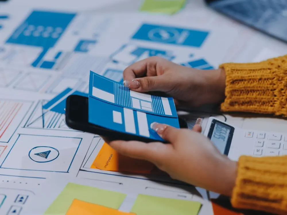 Mans sostenen un esbós d’interfície sobre un mòbil damunt de wireframes impresos